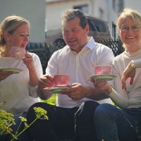 From left to right: Annette Ehrlich, Holger K&ouml;ckritz and Annette Currle sit in the garden and enjoy tea from pink cups. They are wearing white tops and smiling at the camera., &copy; Perle Uhlbach
