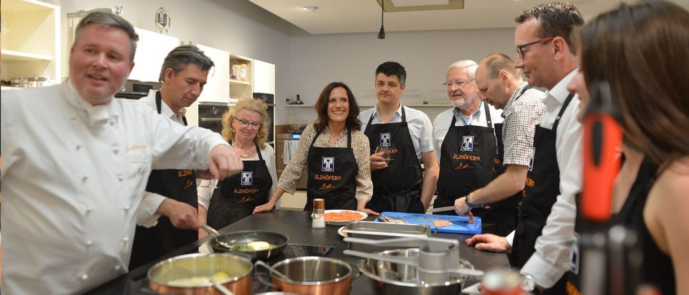 Menschen in einer Küche tragen 'Ilzhöfer's' Schürzen und kochen gemeinsam. Ein Koch zeigt etwas in einer Pfanne., © Ilzhöfer, Stuttgart