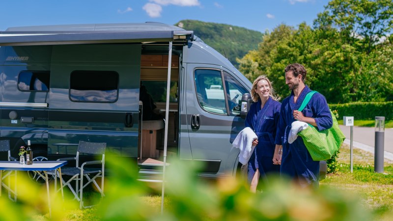 A couple in blue bathrobes is standing, smiling, in front of a modern RV at a sunny campground., &copy; Thomas Niederm&uuml;ller