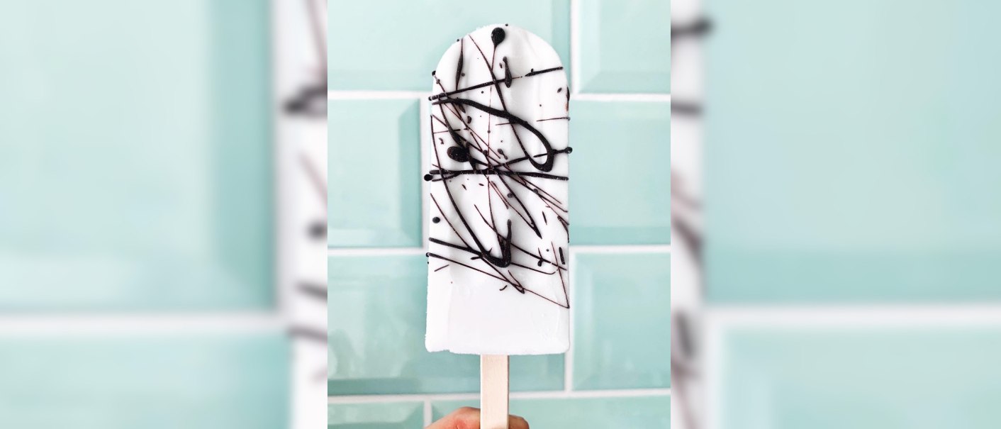 Ein weißes Eis am Stiel mit schwarzer Schokoladendekoration vor einer hellblauen Kachelwand., © Zur Schleckerei Ein weißes Eis am Stiel mit schwarzer Schokoladendekoration vor einer hellblauen Kachelwand., © Zur Schleckerei