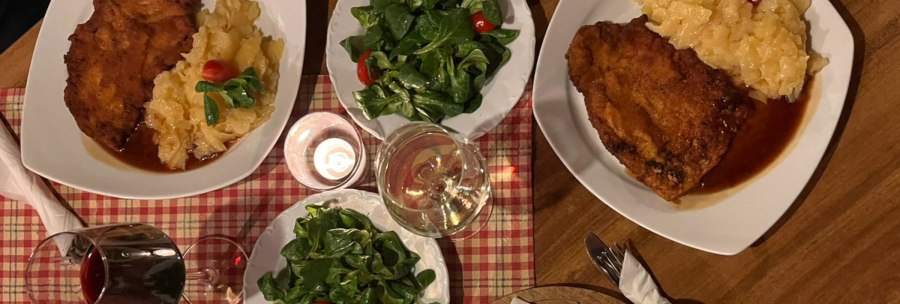 Zwei Teller mit Schnitzel, Kartoffelsalat und grünem Salat. Dazu Rot- und Weißwein auf kariertem Tischtuch., © Weinhof Zaiß