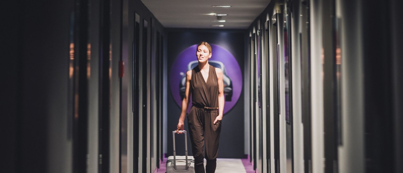 Eine Frau in einem dunklen Outfit zieht einen Koffer durch einen schmalen, modern gestalteten Flur mit lila Akzenten., © Abacco Hotels GmbH Eine Frau in einem dunklen Outfit zieht einen Koffer durch einen schmalen, modern gestalteten Flur mit lila Akzenten., © Abacco Hotels GmbH