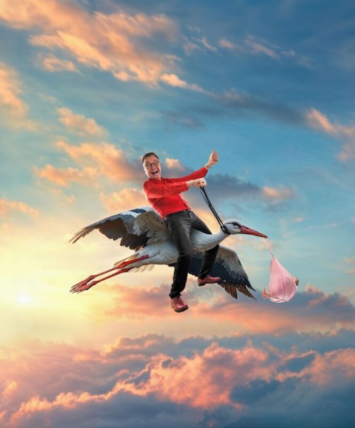 A man in a red shirt rides happily on a stork carrying a bundle in front of a colorful sky with clouds., &copy; Renitenztheater Stuttgart e.V.