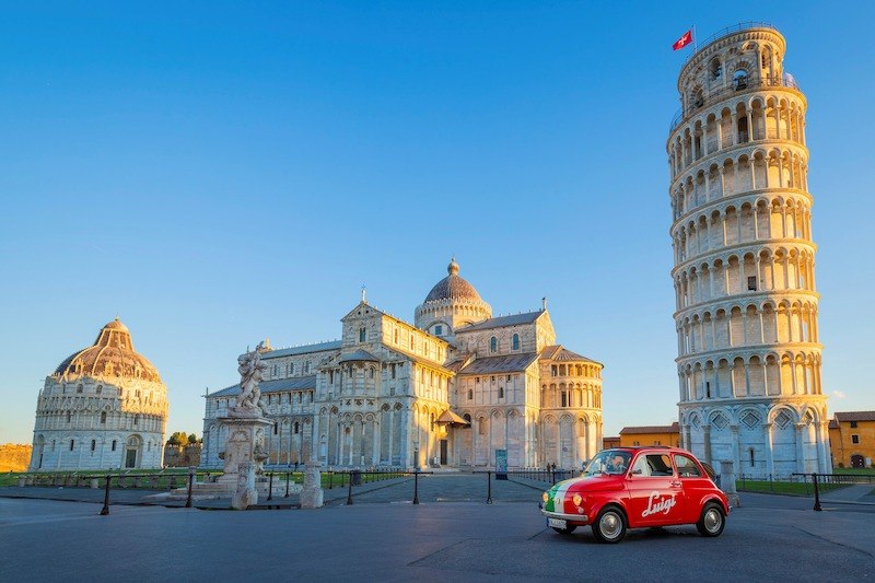 Ein roter Fiat 500 parkt vor dem Schiefen Turm von Pisa und dem Dom, beleuchtet von der Abendsonne., &copy; Theaterhaus Stuttgart e.V.