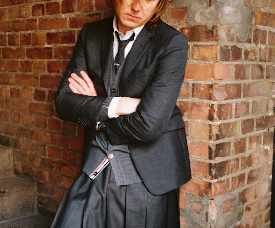 Person in einem dunklen Anzug und Kilt, verschränkt die Arme, steht vor einer Backsteinwand., © C2 Concerts GmbH