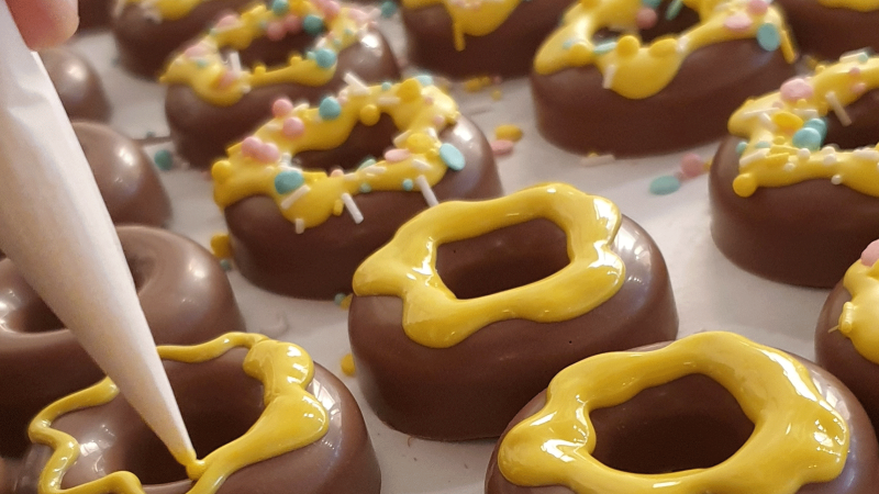 Chocolate rings are decorated with yellow icing and colorful sprinkles. One hand holds a piping bag., © kesselglueck.de Chocolate rings are decorated with yellow icing and colorful sprinkles. One hand holds a piping bag., © kesselglueck.de
