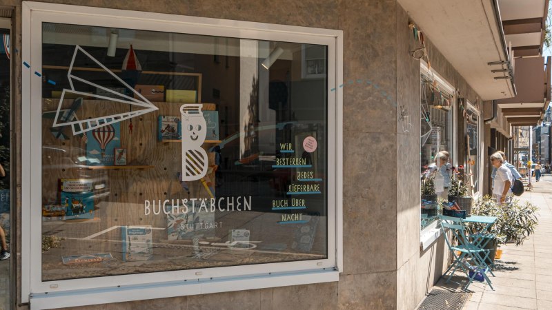 Schaufenster des Buchladens 'Buchstäbchen' in Stuttgart. Dekoriert mit Büchern und einem Papierflieger. Passanten betrachten das Schaufenster., © SMG, Sarah Schmid