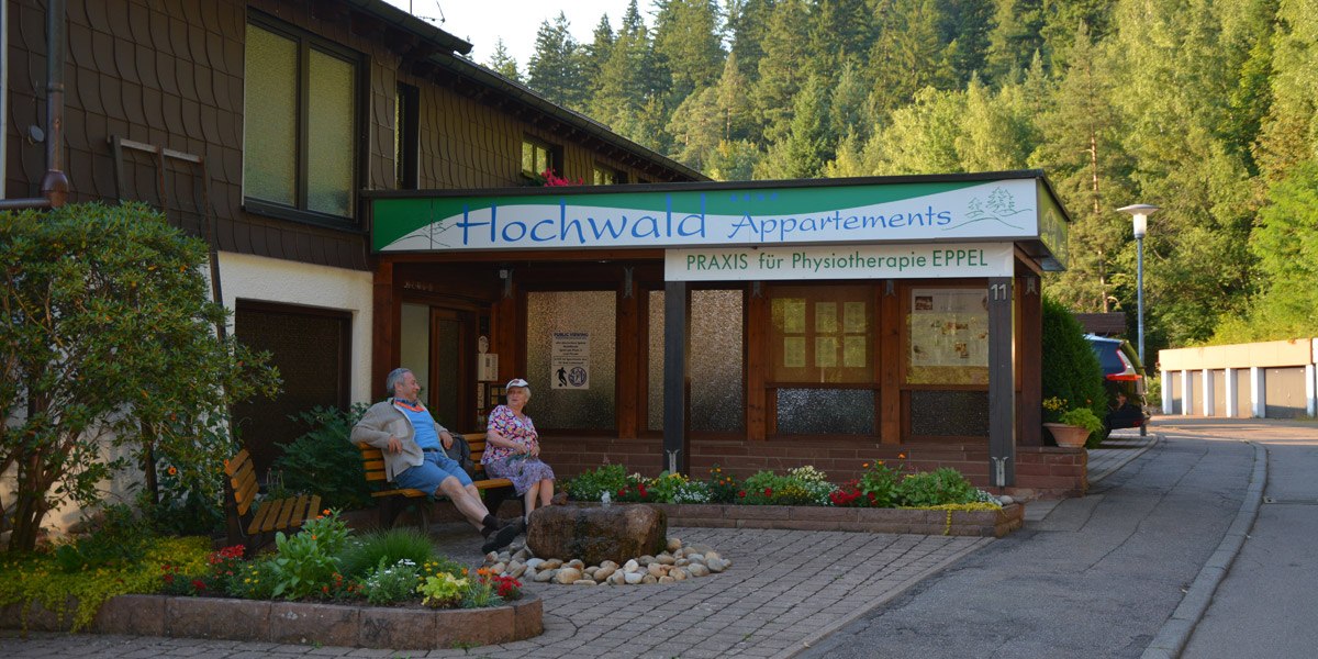 Building with sign 'Hochwald Appartements' and 'Praxis f&uuml;r Physiotherapie EPPEL'. Two people are sitting on a bench, surrounded by plants and trees., &copy; Hotel Hochwald