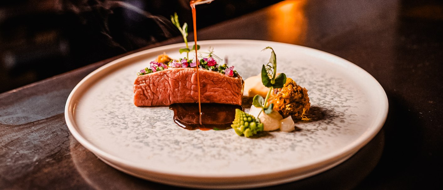 Ein kunstvoll angerichtetes Gourmetgericht mit Fleisch, Gemüse und Sauce auf einem eleganten Teller. Eine Sauce wird darüber gegossen., © wosilatfotografie Ein kunstvoll angerichtetes Gourmetgericht mit Fleisch, Gemüse und Sauce auf einem eleganten Teller. Eine Sauce wird darüber gegossen., © wosilatfotografie