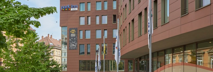 The Park Inn by Radisson Stuttgart has a modern façade with flags in front of it. A tree and a road are visible in the background., © Park Inn by Radisson Stuttgart The Park Inn by Radisson Stuttgart has a modern façade with flags in front of it. A tree and a road are visible in the background., © Park Inn by Radisson Stuttgart