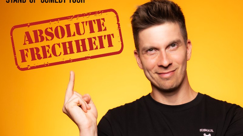 Person in a black T-shirt and blue jeans in front of an orange background, pointing upwards with a finger and smiling slightly., &copy; Renitenztheater Stuttgart e.V.