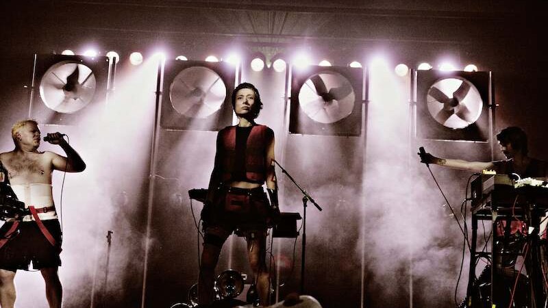 Drei Musiker stehen auf einer Bühne, umgeben von Nebel und großen Ventilatoren im Hintergrund. Einer hält ein Mikrofon, während die anderen an Instrumenten arbeiten., © Theater Rampe Drei Musiker stehen auf einer Bühne, umgeben von Nebel und großen Ventilatoren im Hintergrund. Einer hält ein Mikrofon, während die anderen an Instrumenten arbeiten., © Theater Rampe