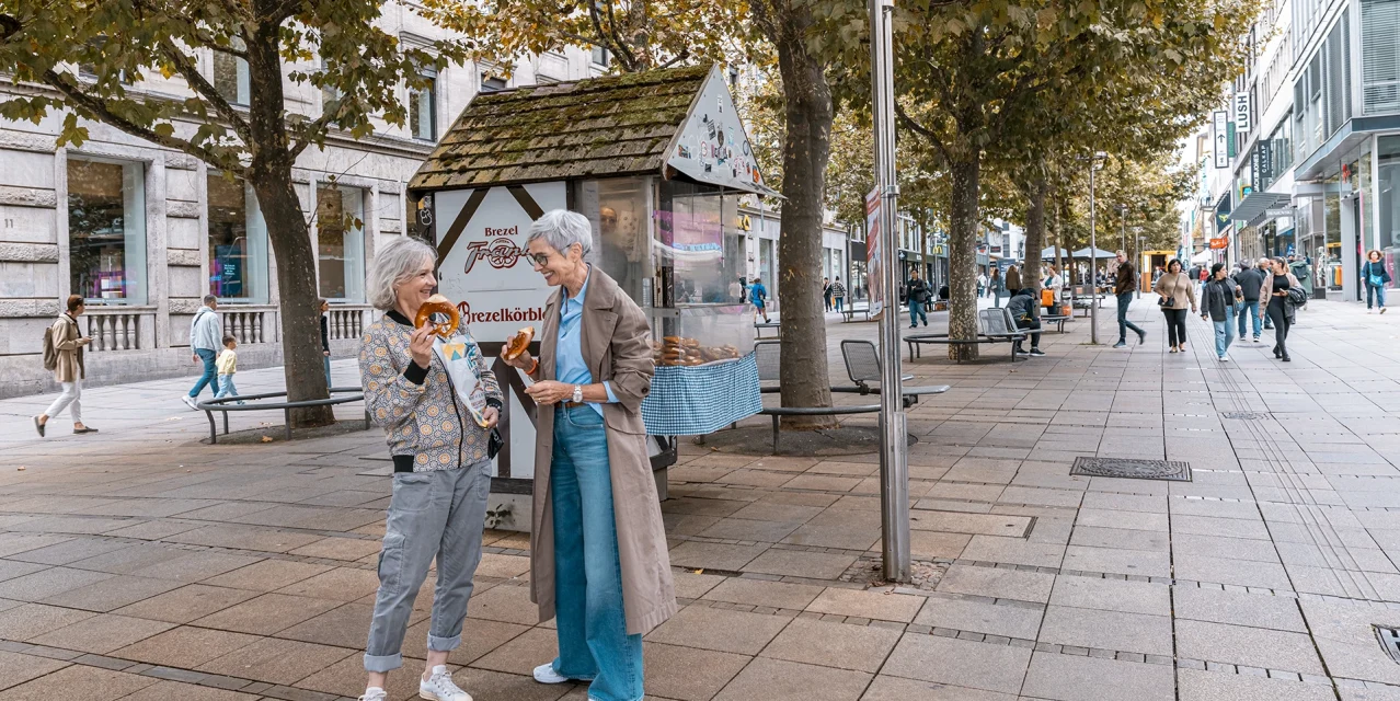 Zwei ältere Frauen genießen Brezeln am Brezelkörble in einer belebten Fußgängerzone., © SMG, Sarah Schmid Zwei ältere Frauen genießen Brezeln am Brezelkörble in einer belebten Fußgängerzone., © SMG, Sarah Schmid