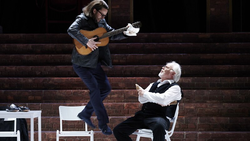 Ein Mann springt mit einer Gitarre in der Hand, während ein anderer auf einem Stuhl sitzt und zuschaut. Die Szene spielt auf einer Bühne mit Backsteinmauer., © Württembergische Staatstheater Stuttgart Ein Mann springt mit einer Gitarre in der Hand, während ein anderer auf einem Stuhl sitzt und zuschaut. Die Szene spielt auf einer Bühne mit Backsteinmauer., © Württembergische Staatstheater Stuttgart