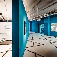 Modern gallery with blue walls, video installations and geometric patterns on the floor. Industrial design with visible ventilation pipes., &copy; Publication only by client - disclosure to third parties only after consultation.