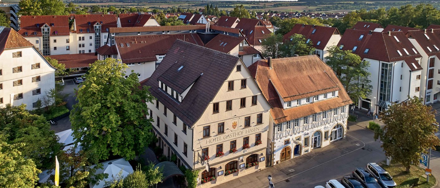 Luftaufnahme eines Hotels mit rotem Dach, umgeben von weiteren Gebäuden und geparkten Autos. Im Hintergrund sind Felder und Hügel zu sehen., © Hotel Gasthof Hasen Luftaufnahme eines Hotels mit rotem Dach, umgeben von weiteren Gebäuden und geparkten Autos. Im Hintergrund sind Felder und Hügel zu sehen., © Hotel Gasthof Hasen