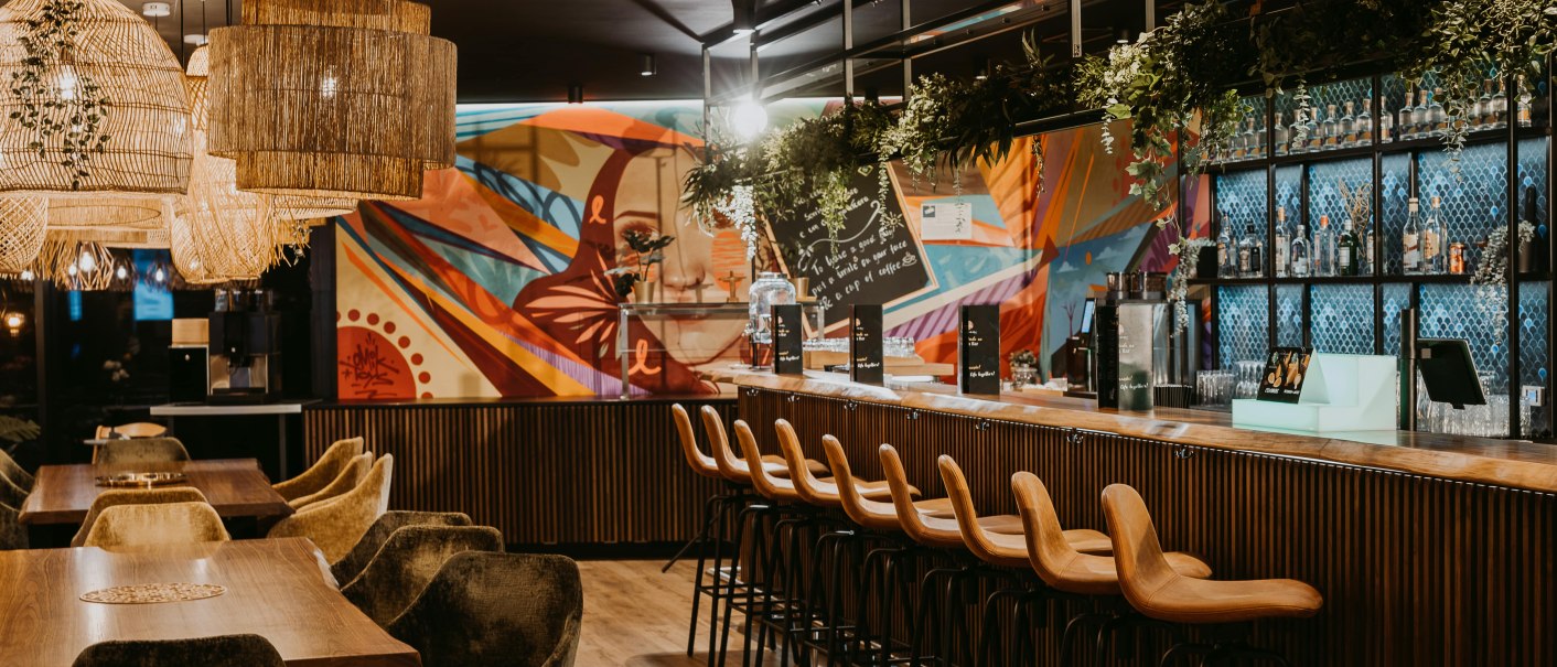 Modern bar with wooden counter, high chairs and colorful wall painting. Hanging lamps and plants decorate the room., © Rioca Stuttgart Posto 4 GmbH Modern bar with wooden counter, high chairs and colorful wall painting. Hanging lamps and plants decorate the room., © Rioca Stuttgart Posto 4 GmbH