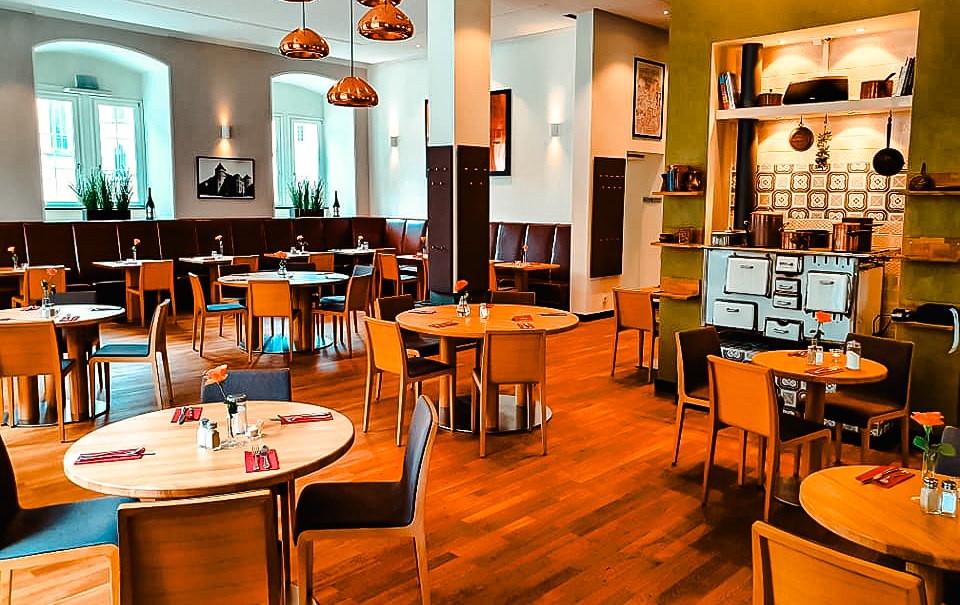 Ein stilvolles Restaurant mit Holztischen und -stühlen, Kupferlampen und einem alten Ofen. Die Atmosphäre ist warm und einladend., © Alte Kanzlei Stuttgart Ein stilvolles Restaurant mit Holztischen und -stühlen, Kupferlampen und einem alten Ofen. Die Atmosphäre ist warm und einladend., © Alte Kanzlei Stuttgart