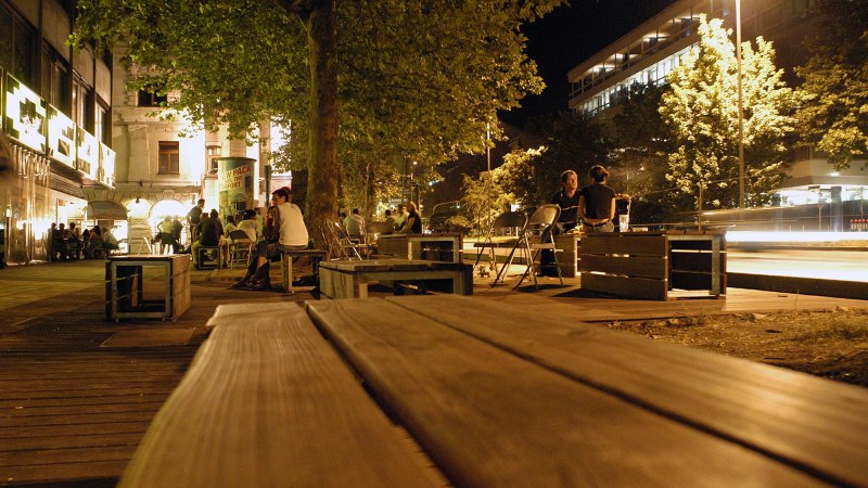 Nachtaufnahme der Theodor-Heuss-Straße in Stuttgart. Menschen sitzen auf Holzbänken unter Bäumen, umgeben von beleuchteten Gebäuden und Straßenlaternen., © Stuttgart-Marketing GmbH, Marc Gilardone Nachtaufnahme der Theodor-Heuss-Straße in Stuttgart. Menschen sitzen auf Holzbänken unter Bäumen, umgeben von beleuchteten Gebäuden und Straßenlaternen., © Stuttgart-Marketing GmbH, Marc Gilardone