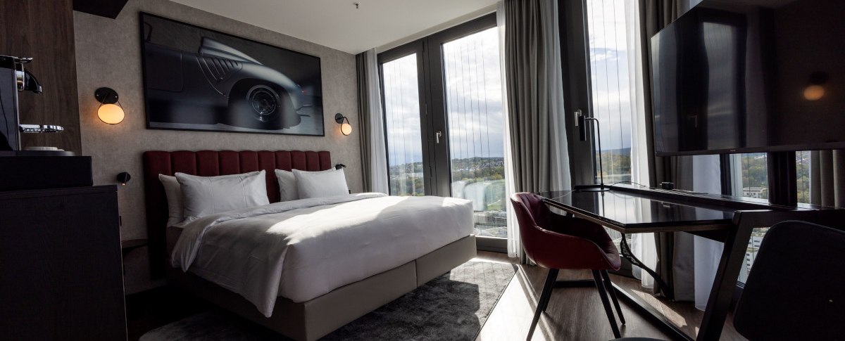 Modernes Eckzimmer mit gro&szlig;em Bett, Schreibtisch, Panoramafenstern und stilvollem Dekor. Ein Bild eines Autos h&auml;ngt an der Wand., &copy; Radisson Hotel Group