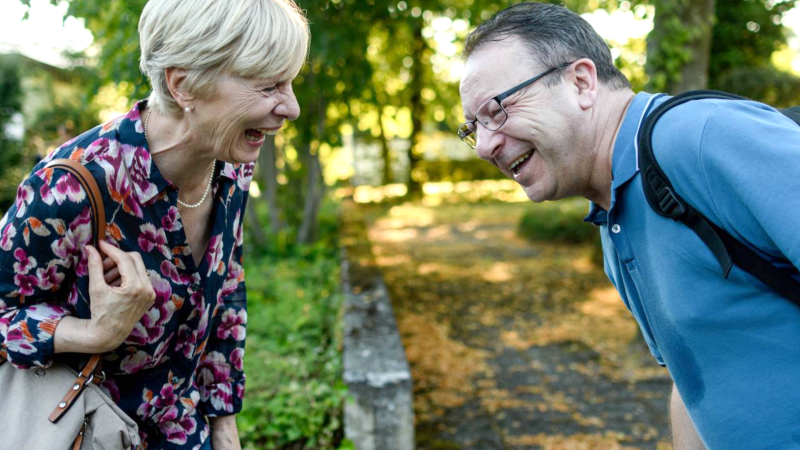 Zwei Personen, eine Frau und ein Mann, lachen herzlich im Freien. Die Frau trägt eine bunte Bluse, der Mann ein blaues Polohemd., © Weltkino