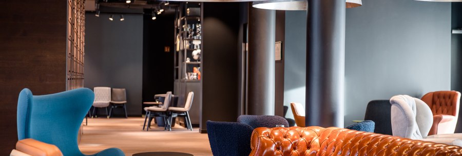 Moderne Lounge mit braunem Ledersofa, blauen und grauen Sesseln, großen Hängelampen und Holzboden. Im Hintergrund sind weitere Sitzgelegenheiten sichtbar., © TOMAS Moderne Lounge mit braunem Ledersofa, blauen und grauen Sesseln, großen Hängelampen und Holzboden. Im Hintergrund sind weitere Sitzgelegenheiten sichtbar., © TOMAS