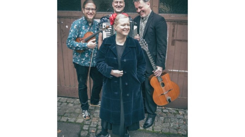 Vier Personen stehen lächelnd mit Musikinstrumenten vor einer Holzwand. Eine Frau trägt einen Mantel, die Männer halten Akkordeon, Geige und Gitarre., © TatjanaCoralieKlytta Vier Personen stehen lächelnd mit Musikinstrumenten vor einer Holzwand. Eine Frau trägt einen Mantel, die Männer halten Akkordeon, Geige und Gitarre., © TatjanaCoralieKlytta