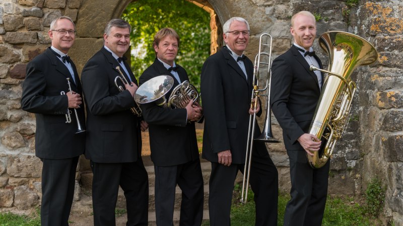 Fünf Männer in Anzügen posieren mit Blechblasinstrumenten vor einer alten Steinmauer mit Torbogen., © SBQ