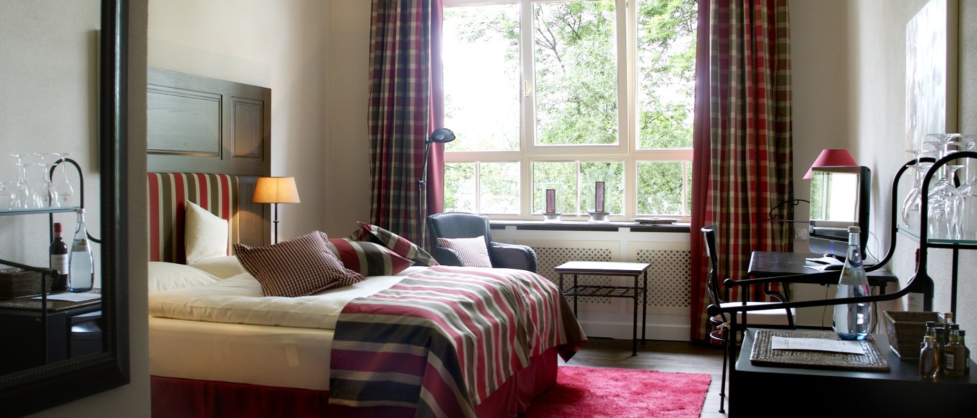 Cozy room with striped bed linen, red carpet and large windows with a view of the greenery. A desk and armchair complete the furnishings., © TOMAS