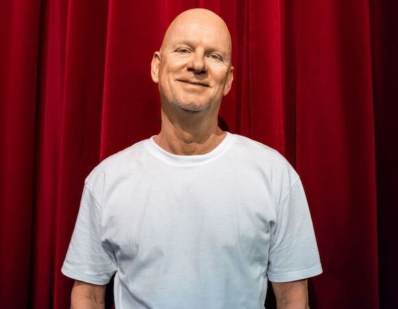 Ein Mann in einem wei&szlig;en T-Shirt steht l&auml;chelnd vor einem roten Vorhang., &copy; ATES Media Group