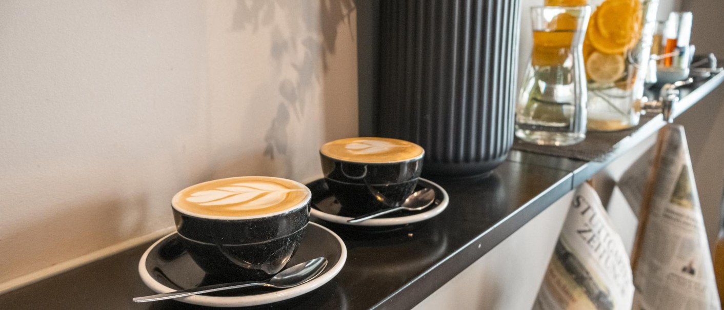 Zwei Cappuccino-Tassen auf einem Regal, daneben Karaffen mit Zitrusfrüchten und Zeitungen. Gemütliche Café-Atmosphäre., © SMG, Sarah Schmid