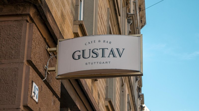 Ein Schild mit der Aufschrift 'Café & Bar Gustav Stuttgart' hängt an einer Hauswand. Der Himmel ist blau., © SMG, Sarah Schmid