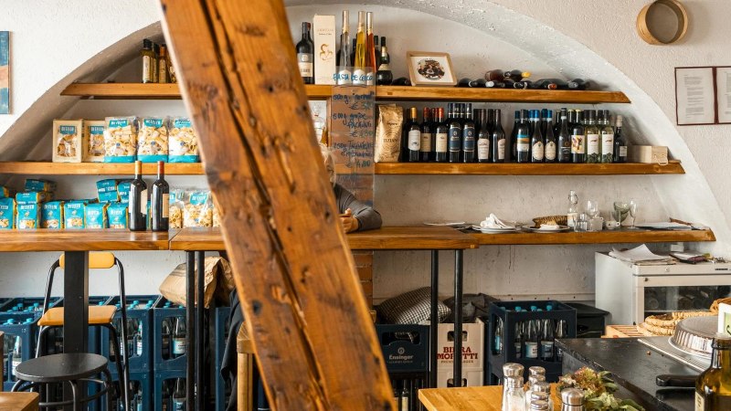 Ein gem&uuml;tlicher Feinkostladen mit Holzregalen, die Weinflaschen und Pasta pr&auml;sentieren. Ein Holzbalken dominiert die Mitte des Bildes., &copy; SMG, Sarah Schmid