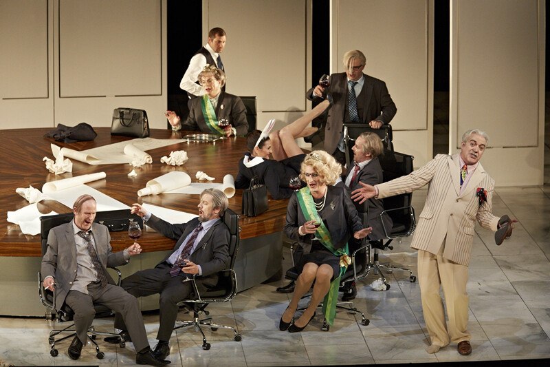 Opernszene mit Darstellern in Anz&uuml;gen und Kost&uuml;men um einen Tisch. Einige sitzen, andere stehen, alle wirken in eine lebhafte Diskussion vertieft., &copy; W&uuml;rttembergische Staatstheater Stuttgart