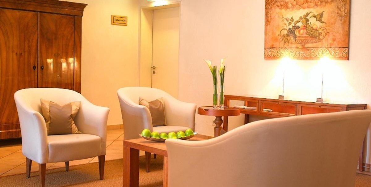 Gemütliche Lobby mit weißen Sesseln, Holztisch mit grünen Äpfeln, Gemälde an der Wand und einem Schrank. Dezente Beleuchtung und Pflanzen., © Hotel am Park Gemütliche Lobby mit weißen Sesseln, Holztisch mit grünen Äpfeln, Gemälde an der Wand und einem Schrank. Dezente Beleuchtung und Pflanzen., © Hotel am Park