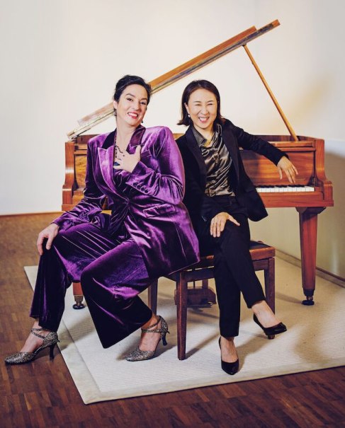 Zwei Frauen in eleganter Kleidung sitzen lächelnd vor einem Klavier. Eine trägt einen lila Anzug, die andere einen schwarzen Anzug., © Wilhelma Theater Zwei Frauen in eleganter Kleidung sitzen lächelnd vor einem Klavier. Eine trägt einen lila Anzug, die andere einen schwarzen Anzug., © Wilhelma Theater