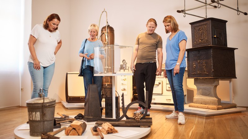 Vier Personen betrachten historische Haushaltsgegenstände im Museum der Alltagskultur. Im Hintergrund sind ein alter Ofen und andere Exponate zu sehen., © FTGRF.de