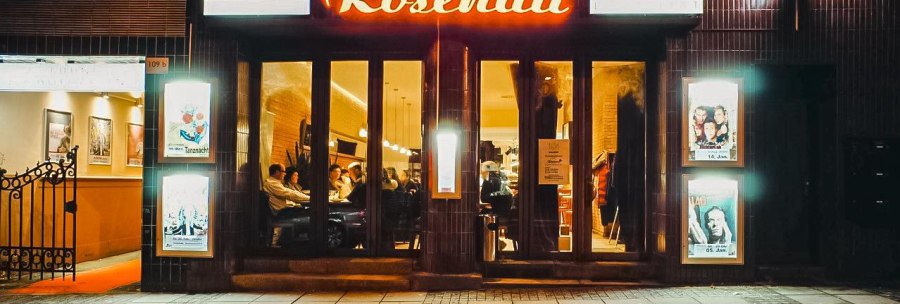 Eingang eines Lokals namens Rosenau mit leuchtendem Schild. Menschen sitzen drinnen, Plakate an den W&auml;nden, gem&uuml;tliche Atmosph&auml;re., &copy; Rosenau e. V., Stuttgart
