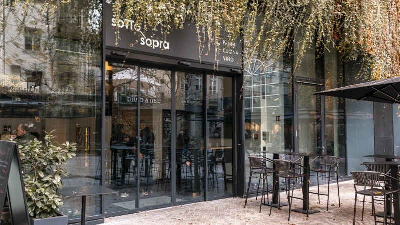 Eingang eines Cafés mit Glasfront und Außenbestuhlung. Pflanzen hängen von oben herab. Menschen sitzen drinnen. Schild mit 'sotto sopra' sichtbar., © SMG, Sarah Schmid