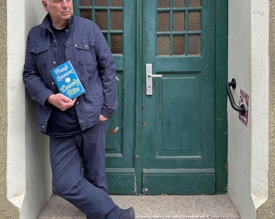 Person lehnt l&auml;ssig an einer gr&uuml;nen T&uuml;r und h&auml;lt ein blaues Buch in der Hand. Die T&uuml;r hat Glasfenster und die Person tr&auml;gt dunkle Kleidung., &copy; Rosenau Kultur e.V.