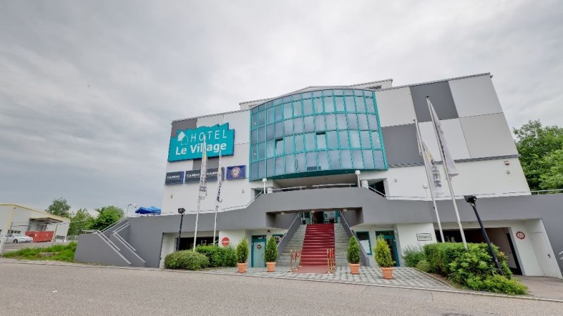 Das Hotel Le Village zeigt eine moderne Fassade mit Glasfront und Treppe. Vor dem Gebäude wehen Fahnen, der Himmel ist bewölkt., © Hotel Le Village
