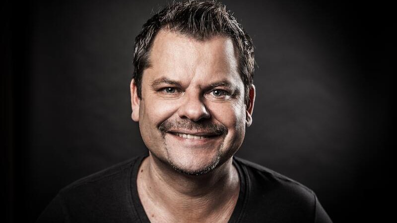 Ein Mann mit kurzem Haar und Bart l&auml;chelt in die Kamera. Der Hintergrund ist dunkel, was den Fokus auf das Gesicht lenkt., &copy; SBEntertainment GmbH & Co.KG