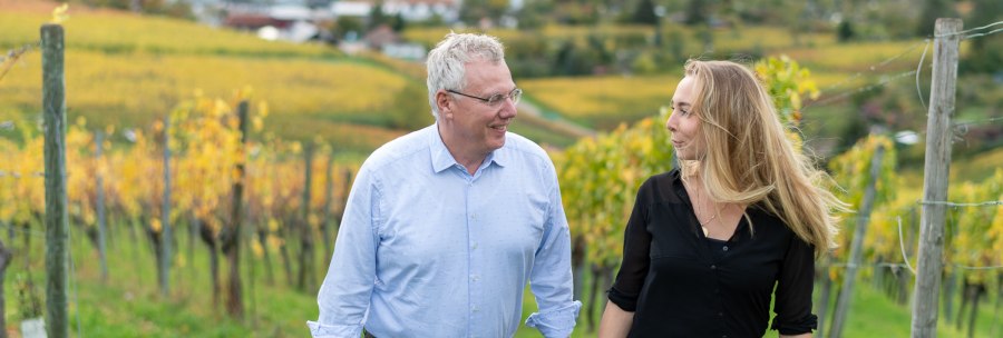 Two people stroll smiling through an autumnal vineyard. Yellow vines and a blurred landscape can be seen in the background., © BURKHARDT HELLWIG