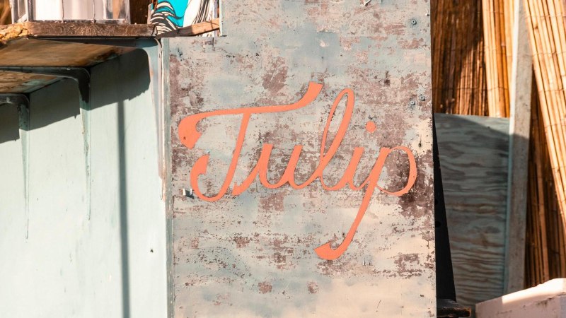 Ein verwittertes Schild mit dem Schriftzug 'Tulip' in orange auf blauem Hintergrund, umgeben von Holzelementen und einem sonnigen Ambiente., © Stuttgart-Marketing GmbH, Sarah Schmid Ein verwittertes Schild mit dem Schriftzug 'Tulip' in orange auf blauem Hintergrund, umgeben von Holzelementen und einem sonnigen Ambiente., © Stuttgart-Marketing GmbH, Sarah Schmid