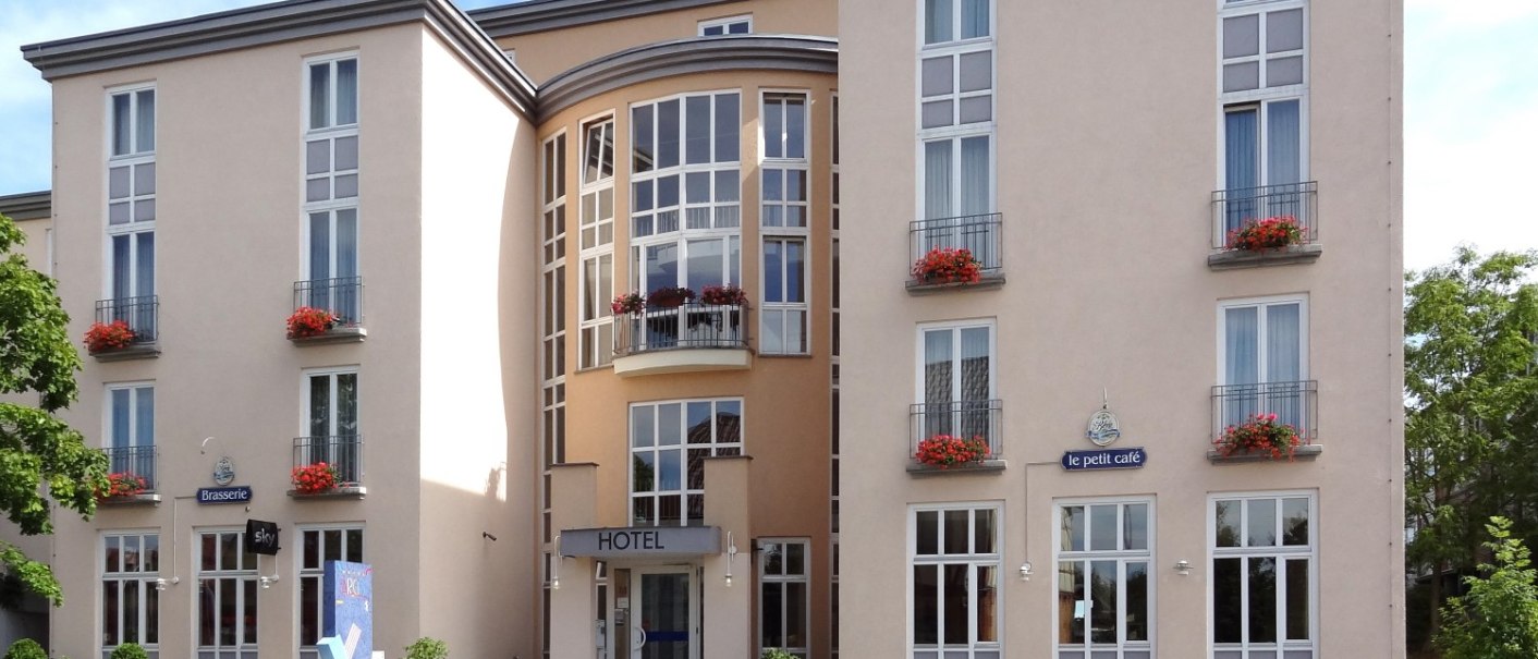 Modernes Hotelgebäude mit großen Fenstern, Balkonen mit roten Blumen und einem blauen "HOTEL"-Schild auf dem Dach., © Hotel Arcis