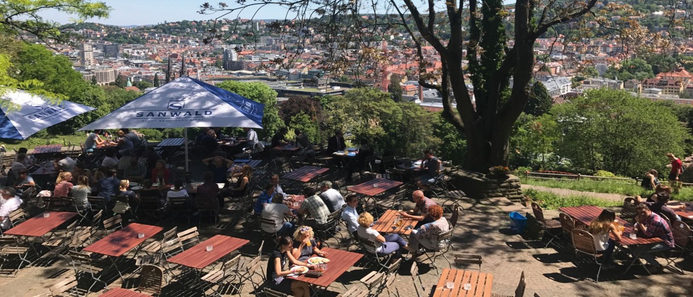 A beer garden on the Karlshöhe with a view of Stuttgart. People sit at tables under parasols, surrounded by trees and a view of the city., © Stuttgart-Marketing GmbH