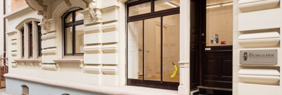 Eingang einer Galerie mit dem Schild 'Die Raumgalerie'. Eine Banane ist an der Glastür angebracht. Das Gebäude hat eine verzierte Fassade., © Dirk Wilhelmy Fotografie