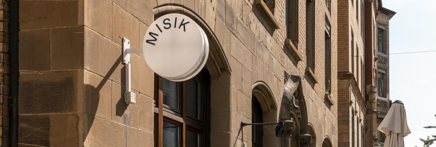 Geb&auml;ude mit Sandsteinfassade, rundes Schild mit 'MISIK', Fenster mit Pflanzen, geschlossene Sonnenschirme auf der Stra&szlig;e., &copy; Stuttgart-Marketing GmbH, Sarah Schmid
