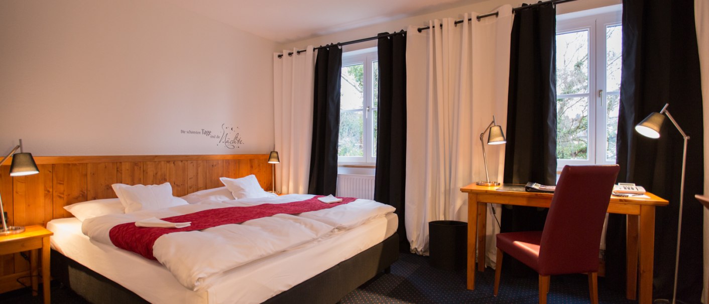 A cozy hotel room with a double bed, a red bedspread, a desk, and a chair. Large windows with black and white curtains., &copy; Hotel B&uuml;rkle