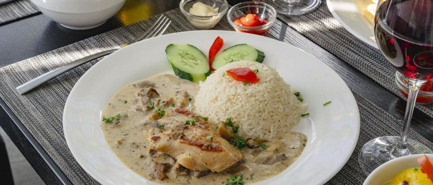 Ein Teller mit Reis, Hähnchen in Sahnesoße, Gurken und Paprika. Daneben ein Glas Rotwein und Salat auf einem gedeckten Tisch., © Plaza INN Stuttgart-Filderstadt Ein Teller mit Reis, Hähnchen in Sahnesoße, Gurken und Paprika. Daneben ein Glas Rotwein und Salat auf einem gedeckten Tisch., © Plaza INN Stuttgart-Filderstadt